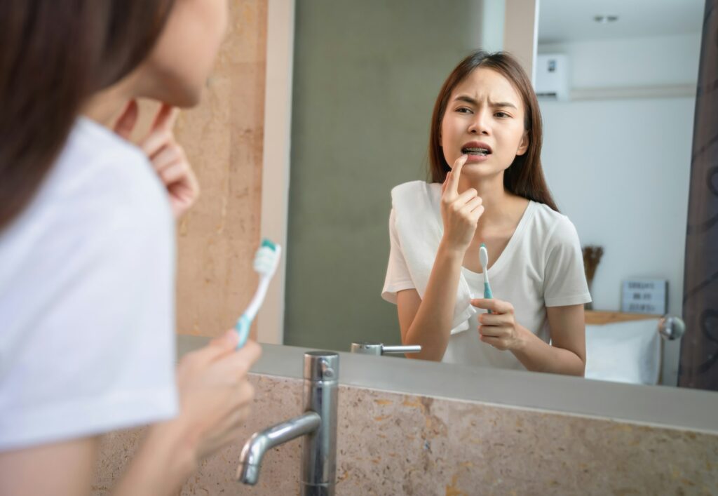 Young girl brushing her teeth in pain, touching a sensitive tooth in the bathroom mirror, highlighting the importance of sensitive teeth treatment at Kids Smiles Pediatric Dentistry in St. Louis, MO